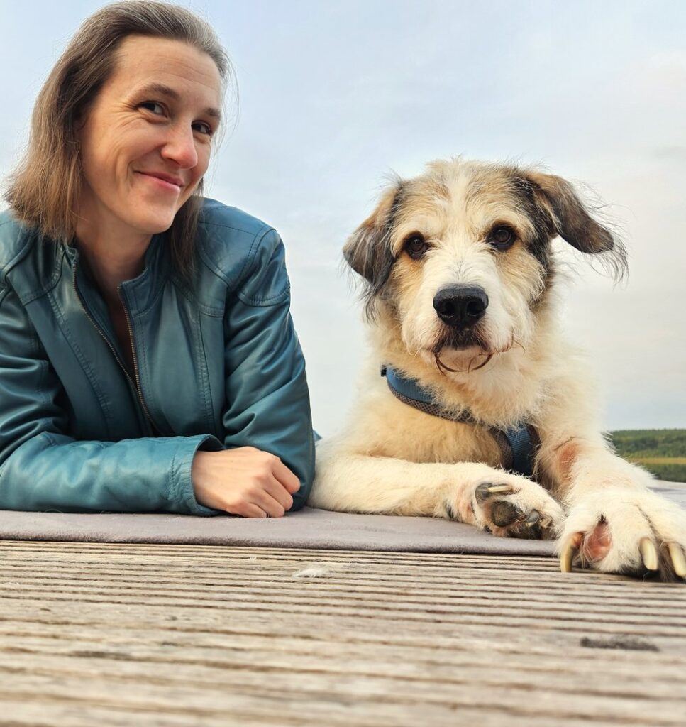 Susanne Krämer liegt lächelnd mit ihrem Hund Socke auf einem Steg am See